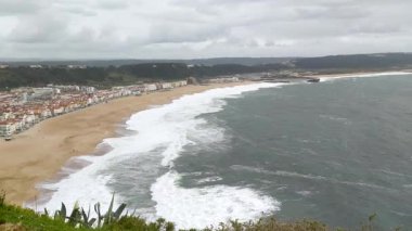Dramatik kıyı manzarası kasvetli bir gökyüzünün altındaki tuhaf bir kasabanın yakınındaki kumlu bir sahile çarpan güçlü dalgaları sergiliyor. Doğa kuvvetleri, seyahat veya uzak konumları çağrıştırır.