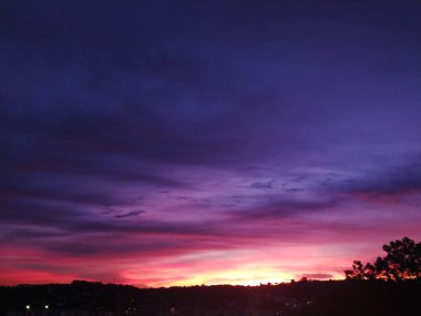 Siluetli bir şehir manzarasında dramatik bir gün batımı. Canlı pembe, mor ve turuncu renkler gökyüzünü boyuyor, nefes kesici, atmosferik bir sahne yaratıyor seyahat, doğa veya karman çorman projeler için mükemmel..