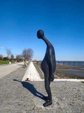 Deniz kenarındaki bir gezinti alanında uzun bir gölge oluşturan soyut bronz heykel. Şekiller, açık mavi gökyüzüne karşı derin düşüncelere ya da yalnızlığa yol açar..