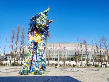 Geri dönüşümlü malzemelerden yapılan renkli vaşak heykeli, berrak mavi gökyüzünün altındaki modern bir spor sahasının önünde görkemli bir şekilde duruyor. Çevre, sanat ve seyahat temaları için ideal.