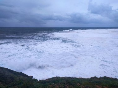 Güçlü dalgaların fırtınalı bir gökyüzünün altında engebeli bir kıyı şeridine çarptığı dramatik deniz manzarası. Doğa, esneklik, sükunet temalarını çağrıştırıyor. Seyahat, hava durumu veya ruh hali görüntüleri için ideal.