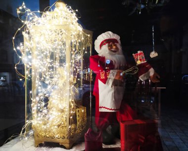 ALCOBACA, PORTUGAL - 30 Aralık 2024: Kutlama penceresi Noel Baba şefini çırpıcı ve ışıklarla antik fenerin yanında hediyelerle sergiliyor. Tatiller, yemek pişirme ve kutlama temaları.