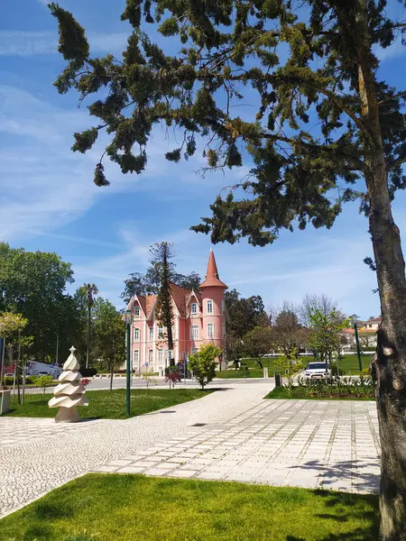 ALCOBACA, PORTUGAL - 27 Nisan 2025: kuleye sahip antik, peri masalı gibi pembe bir bina. Paved Plaza, çimenler ve ağaçlar. Güneşli bir gün, seyahat ya da mimari tema.
