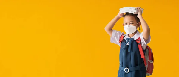Back to school banner idea concept, Asian school girl wearing medical face mask with hold books on her head, isolated on yellow background with Clipping paths for design work empty free space