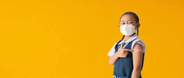 Back to school banner idea concept, Asian student little girl showing pointing with finger to bandage on her arm shoulder wearing protective mask done with vaccinations
