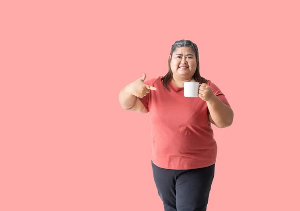 Fat woman asian happy smiling holding a mug with hand pointing, isolated on pink  background