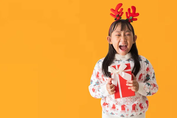 喜庆的亚洲小女孩穿着一件圣诞毛衣 头戴驯鹿角 抱着圣诞礼盒笑得开心 新年快乐 与白色背景隔离 图库照片