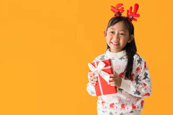 喜庆的亚洲小女孩穿着一件圣诞毛衣 头戴驯鹿角 抱着圣诞礼盒笑得开心 新年快乐 与白色背景隔离 图库图片