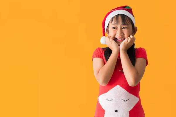 穿着红色圣诞服装戴着圣诞帽的亚洲小女孩快乐地微笑着 站在那里摆出一副奇怪的姿势 孤零零地躺在黄色的背景上 图库图片