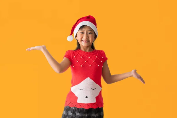 身穿红色圣诞服装戴着圣诞帽的亚洲小女孩快乐地微笑着 站在那里摆出一副奇怪的姿势 孤零零地躺在黄色的背景上 免版税图库图片