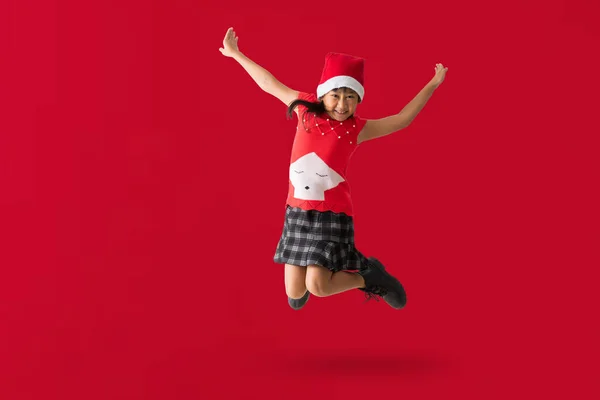 身穿红色圣诞服装 头戴圣诞帽 快乐地微笑着的亚洲小女孩 欢欢喜喜地跳舞 跳跃着 全身上下都挂着一幅红色背景的照片 免版税图库照片