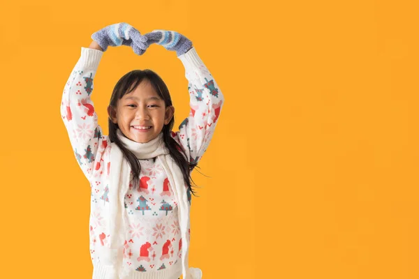 穿着圣诞羊毛衫服装的快乐的亚洲小女孩的画像 戴着手工制作的心形手套 孤立在黄色背景下 免版税图库照片
