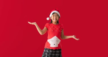 Noel Baba şapkalı mutlu Asyalı küçük bir kızın portresi. El ele tutuşmuş, kırmızı arka planda yalnız poz veriyor.