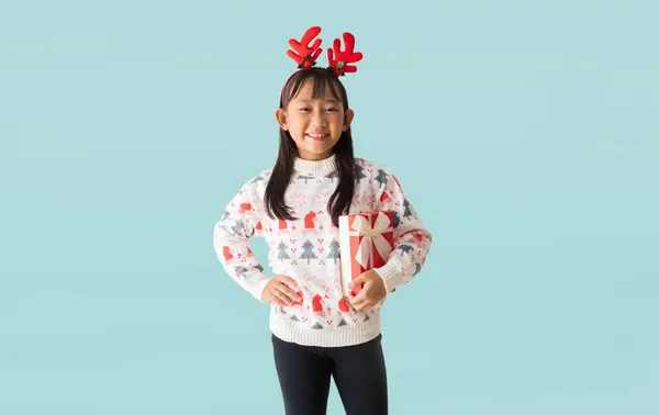 穿着圣诞羊毛衫的快乐的亚洲小女孩的画像 手牵着圣诞礼品盒 新年快乐 孤零零地躺在苍白浅蓝色的背景上 图库图片