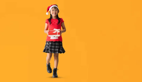 笑容满面的亚洲小女孩戴着圣诞礼帽手握着圣诞礼盒 新年快乐 全身上下被黄色的背景隔开 免版税图库照片