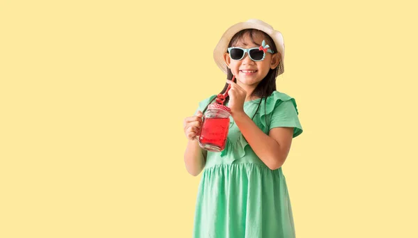 赤い甘い水のガラスを保持する帽子とサングラスを身に着けているハッピーアジアの少女 パステル黄色の背景に隔離された休日の夏のファッショングリーンドレス ストックフォト