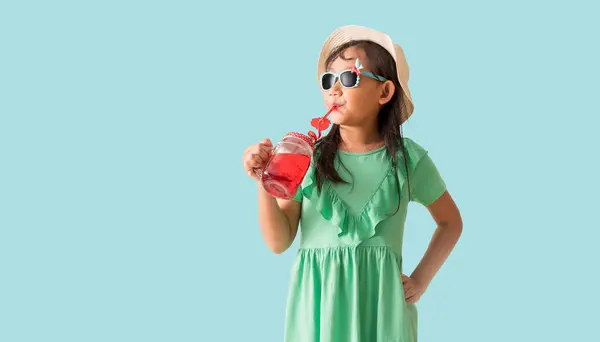 赤い甘い水のガラスを保持する帽子とサングラスを身に着けている幸せなアジアの少女 パステルブルーの背景に隔離されたホリデー夏のファッショングリーンドレス ロイヤリティフリーのストック写真