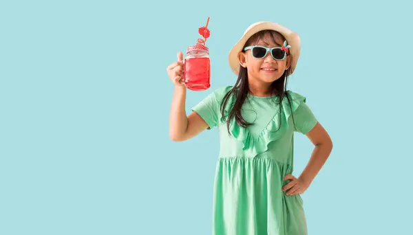 赤い甘い水のガラスを保持する帽子とサングラスを身に着けている幸せなアジアの少女 パステルブルーの背景に隔離されたホリデー夏のファッショングリーンドレス ストック画像