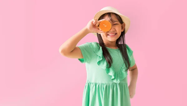 ハッピーアジアの少女は 顔にオレンジ色のスライスを保持するサングラスで帽子をかぶる パステルピンク色の背景に孤立した休日の夏のファッショングリーンドレス ストック写真