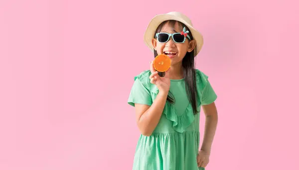 快乐的亚洲小女孩 头戴一顶戴着太阳镜的帽子 手里拿着桔子片 穿着夏令时尚的绿色连衣裙 背景是淡淡的粉色 图库照片