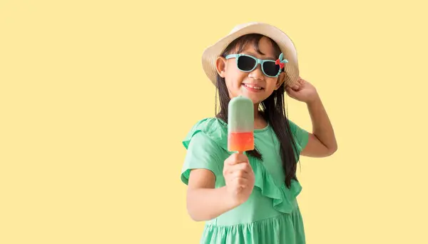 ハッピーアジアの少女は アイスクリームを保持するサングラスで帽子をかぶる パステル黄色の背景に隔離されたホリデー夏のファッショングリーンドレス ストック画像