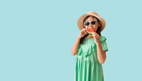快乐的亚洲小女孩 头戴一顶戴着太阳镜的帽子 手里拿着西瓜片 穿着淡蓝色背景的夏日时尚服饰 免版税图库照片