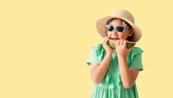 快乐的亚洲小女孩 头戴一顶戴着太阳镜的帽子 手里拿着西瓜片 穿着淡黄相间的夏装 图库照片