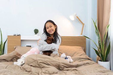 Mutlu gülümseyen Asyalı Taylandlı kız. Yatakta oturan oyuncak ayıyı kucakla. Sıcak ve rahat bir atmosfer.