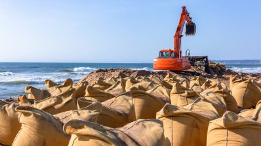 Installment construction with excavator industrial machine on beach coastline of large canvas sand bags placed as a protection barrier wall from ocean sea water storms and tidal surges . clipart