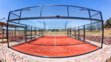 Padel Tennis game on red astro surface with glass walled enclosed court  in countryside on a blue sky day. clipart