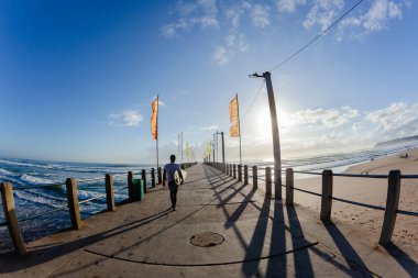 İskele platformunda sörf tahtasıyla yürüyen genç sörfçü. Arkasında tanınmaz hale gelmiş, tanınmaz hale gelmiş bir adam. Okyanus girişi iskelede sörf yapmak için..