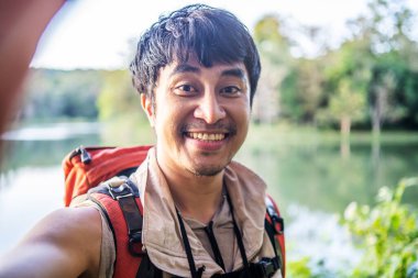 Genç Asyalı çekici erkek turistler ellerinde kameralı cep telefonuyla yeşil yaz çimlerinde selfie çekiyorlar ve kameraya bakıp güzelliğe hayran kalıyorlar.