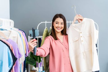 Moda mağazasında genç Asyalı kadın işyeri sahibi canlı yayında moda kıyafetleri satıyor. Sosyal medyada sunum için blog eleştirmeni kıyafetleri. Çevrimiçi alışveriş kavramı.