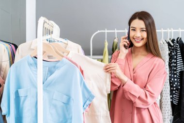 Güzel Asyalı kadın çevrimiçi satıcı müşterinin siparişlerini onaylıyor. E-ticaret işyeri sahibi mağazada telefonla konuşarak kıyafet seçiyor ve elbise seçiyor..