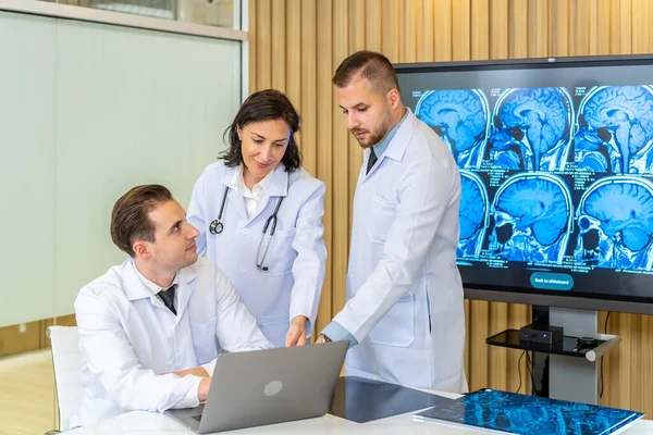 Team of three doctors discusses the results of an MRI scan of the patient\'s head in the consulting room of modern hospital, physician discussing about treatment for patient looking in laptop