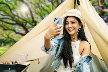 Asyalı bir kadın, macera ve aktif seyahat için ulusal parkta kamp yaparken cep telefonu kullanıyor ve çadırında selfie çekiyor.