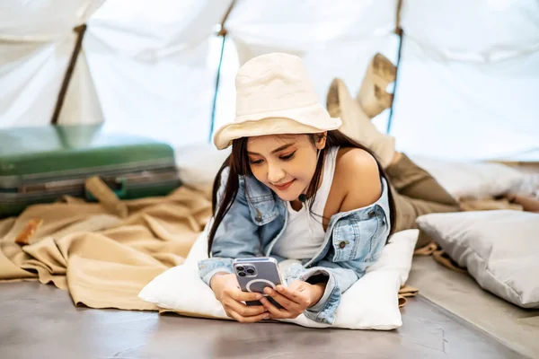 Asian woman is using mobile technology phone with internet connection at her tent while camping outdoor during summer time in national park for adventure and active travel