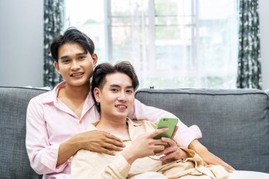 Sevimli erkek eşcinsel çift evde vakit geçiriyor. Genç mutlu lgbtq çifti kanepeye uzanıyor ve Smartphone kullanıyor, ekommerce uygulamasında online alışveriş yapıyor, sosyal medyayı birlikte kontrol ederken eğleniyor.