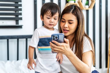 Genç Asyalı annesi cep telefonunu kullanıp ekranda komik sosyal medya videoları izleyerek evde yatakta rahatça alışveriş yapıyor.