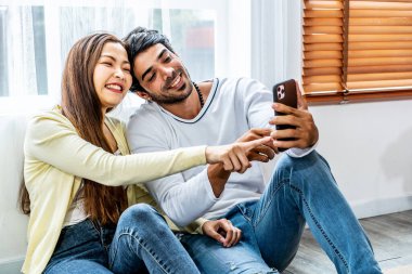 Rahatlamış genç bir çift ya da arkadaşlar evdeki oturma odasında cep telefonuyla video konferansında konuşarak akıllı telefonu işaret eden mutlu genç adam ve kadın.