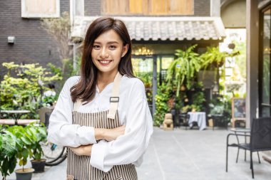 Başarılı küçük işletmeler sahibi genç Asyalı kadın kameraya bakıyor çapraz kollar pozu veriyor, önlük giymiş garsonun portresi iş yerinde arkadaşça gülümsüyor.