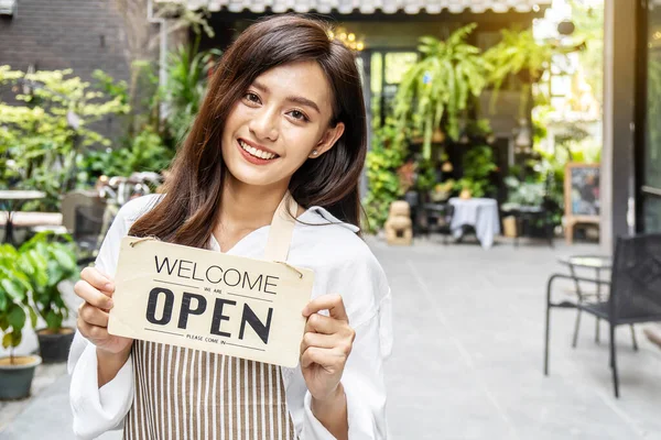 Business owner happy beautiful young asian woman in apron looking at camera, we\'re open sign on front door smiling welcoming clients to new cafe, People and start-up concept.