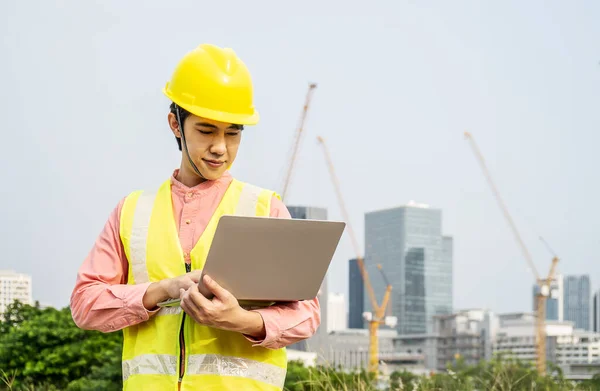 Engineer or construction worker Asian man is holding the laptop and working concentrated at building plan, Hard hat help protect with operation