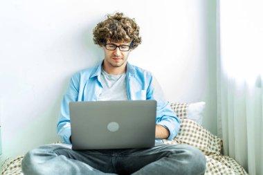 Evden çalışıyorum. Bilgisayarda çalışan beyaz bir genç adam. Çevrimiçi dinleme öğrenim kursu konferansı çağrısı. Yatakta uzanmak konsepti, evde serbest çalışmak.