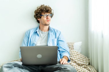 Evden çalışıyorum. Genç, beyaz bir adam bilgisayar laptopunda çalışıyor ve başka yere bakıyor, çevrimiçi öğrenim kurslarını dinliyor eğitim konferanslarını dinliyor yatağa uzanıyor, evde serbest çalışıyor.