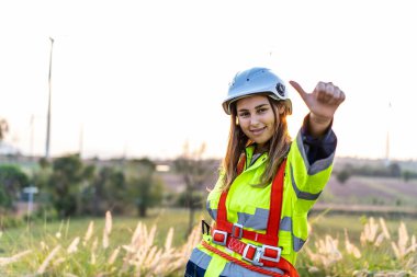 Üniformalı, gülümseyen beyaz kadın mühendis rüzgar türbini çiftliğinde başarılı bir projeyi kutlarken mutlu bir şekilde başparmak kaldırıyor. Mutlu işçi işi seviyor.