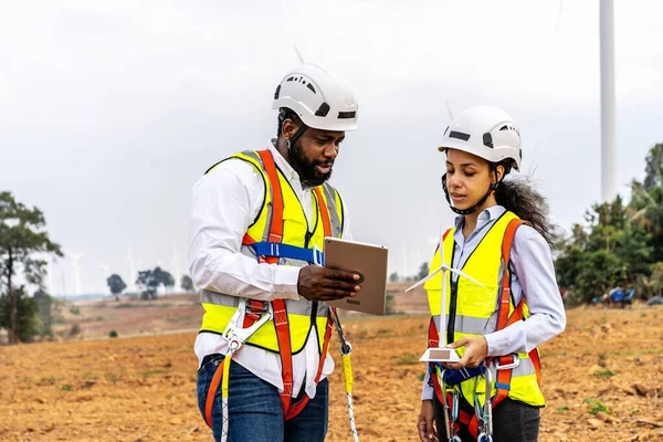 Front view of two engineers african american man and woman in uniform discuss and use tablet working with wind turbine model at wind turbine farm, renewable energy, technology, power, service