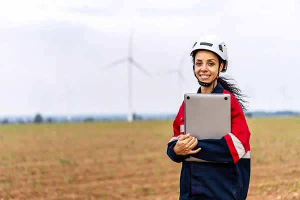 A portrait of smiling engineer african american woman in uniform working in the wind turbine area, and look at camera confidently away from the white windmill