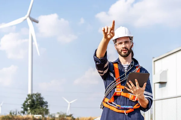 Portrait of caucasian engineer in uniform wearing helmet, viewing data on tablet about system installation in the background wind turbine. business success, wind turbine generate electricity