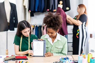 Genç iki moda tasarımcısı, grafik tasarımcısıyla dijital tablet toplantısını kullanarak dikiş atölyesinde çalışırken ekranda yeni elbise tasarımı koleksiyonunu tartışıyor.